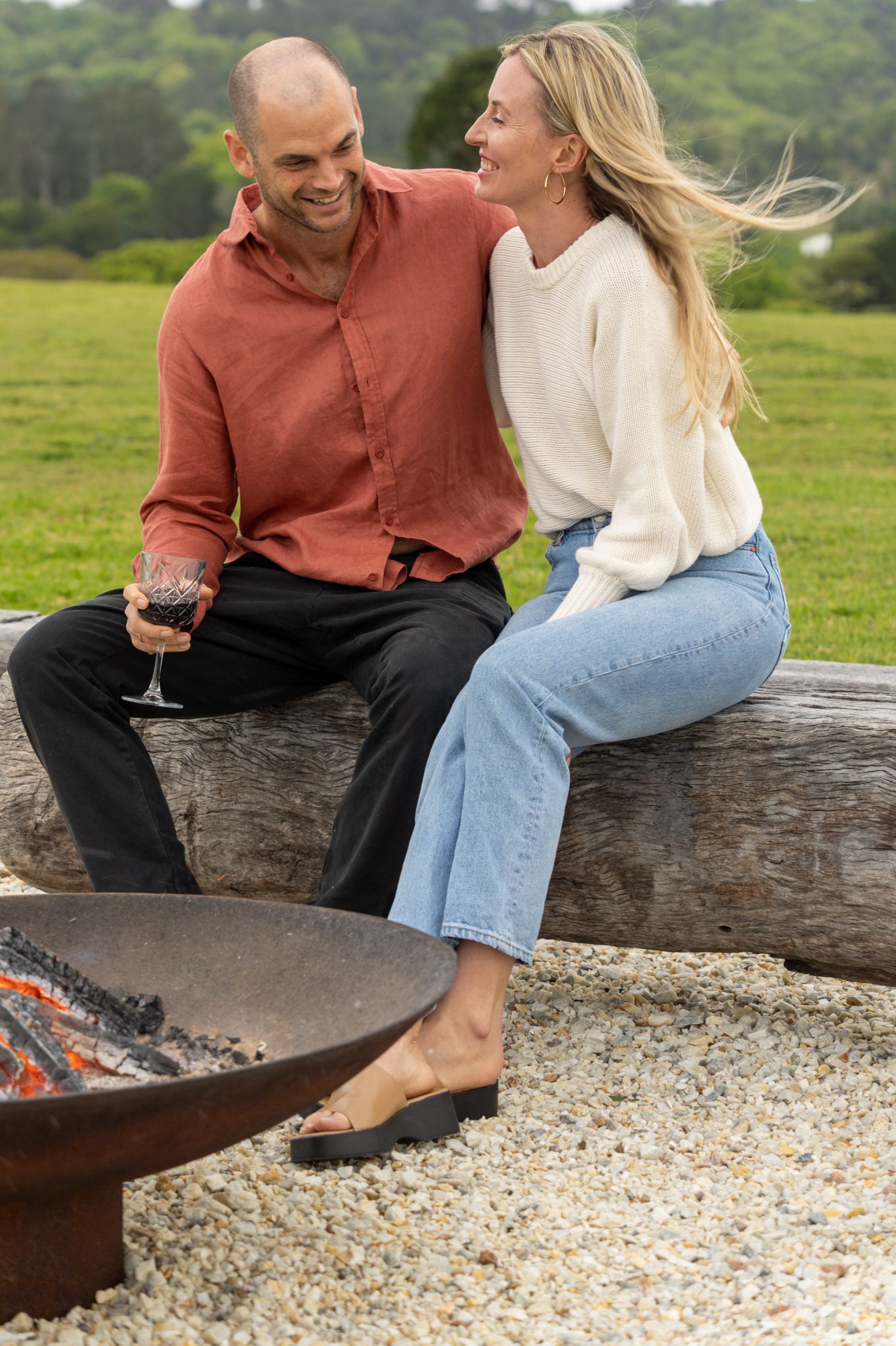 casually dressed man and woman sitting on a wooden bench drinking red wine and smiling at each other by the heartwood farm cottage fire pit