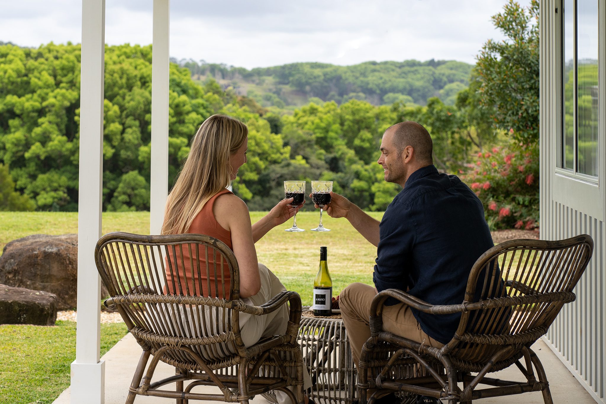 woman and man sitting on the heartwood farm cottage veranda toasting their red wine glasses with the byron bay hinterland in the background