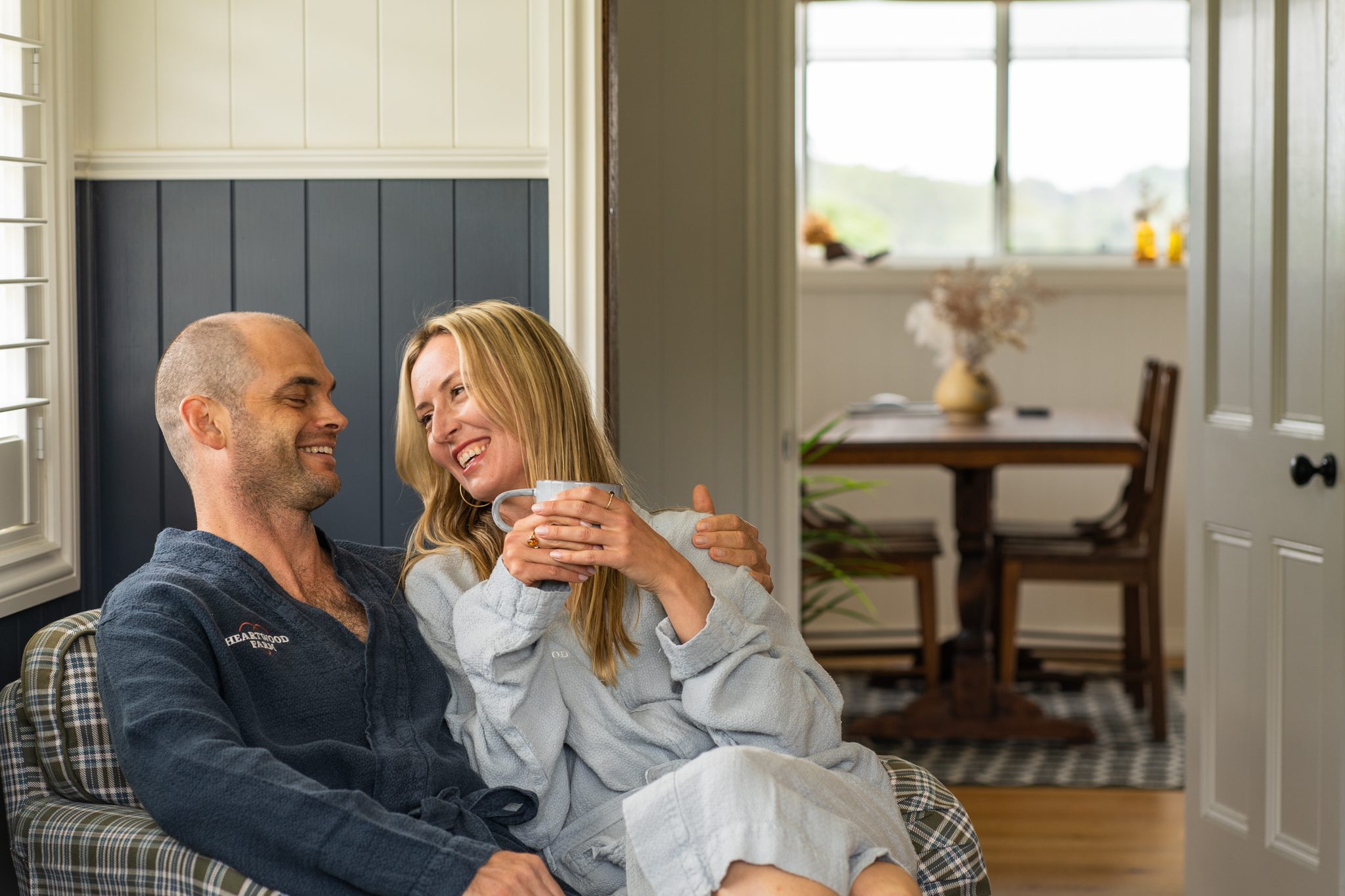 bald man and blonde woman wearing heartwood farm cottage blue dressing gowns sitting on the same bedroom chair smiling and relaxing with each other