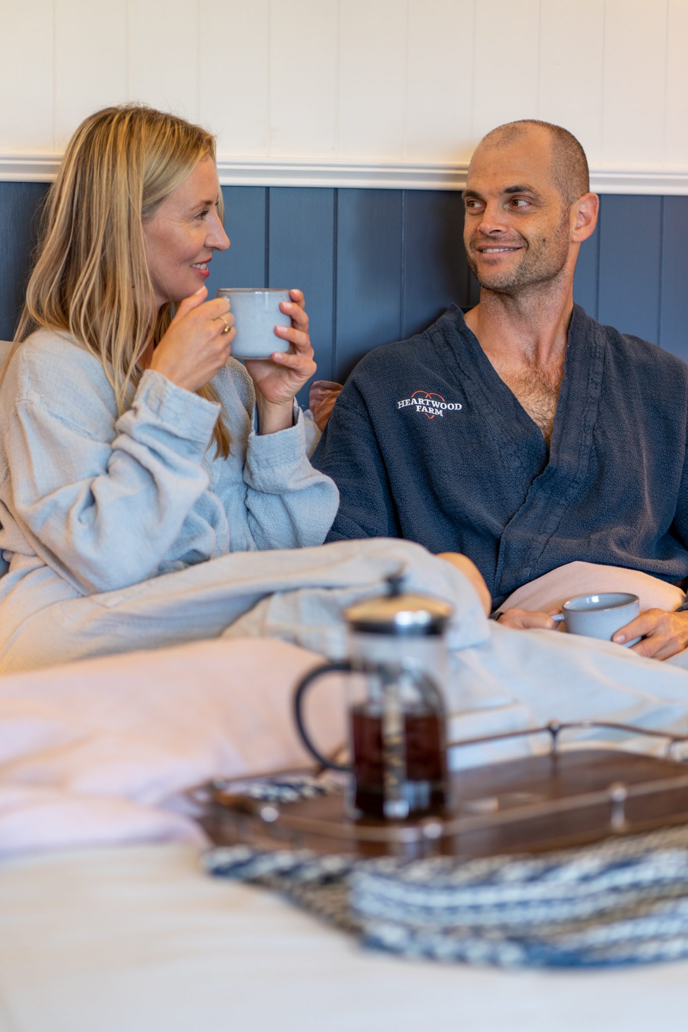 man and woman wearing heartwood farm blue dressing gowns sitting in bed and smiling at each other drinking coffee at heartwood farm cottage
