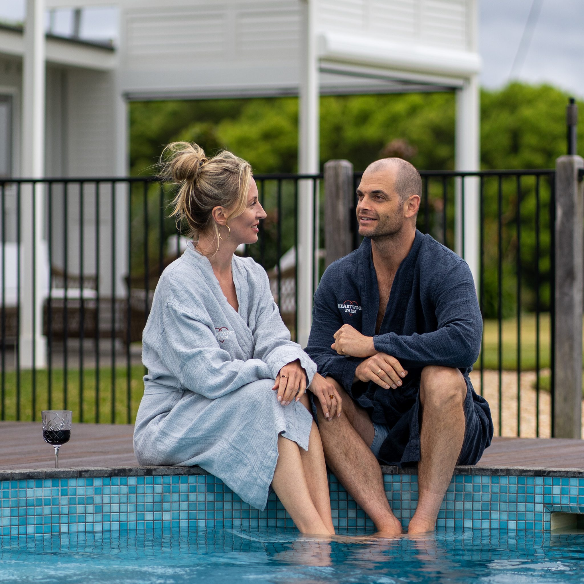 bald man and blonde women sitting by the heartwood farm cottage pool in their dressing gowns drinking red wine