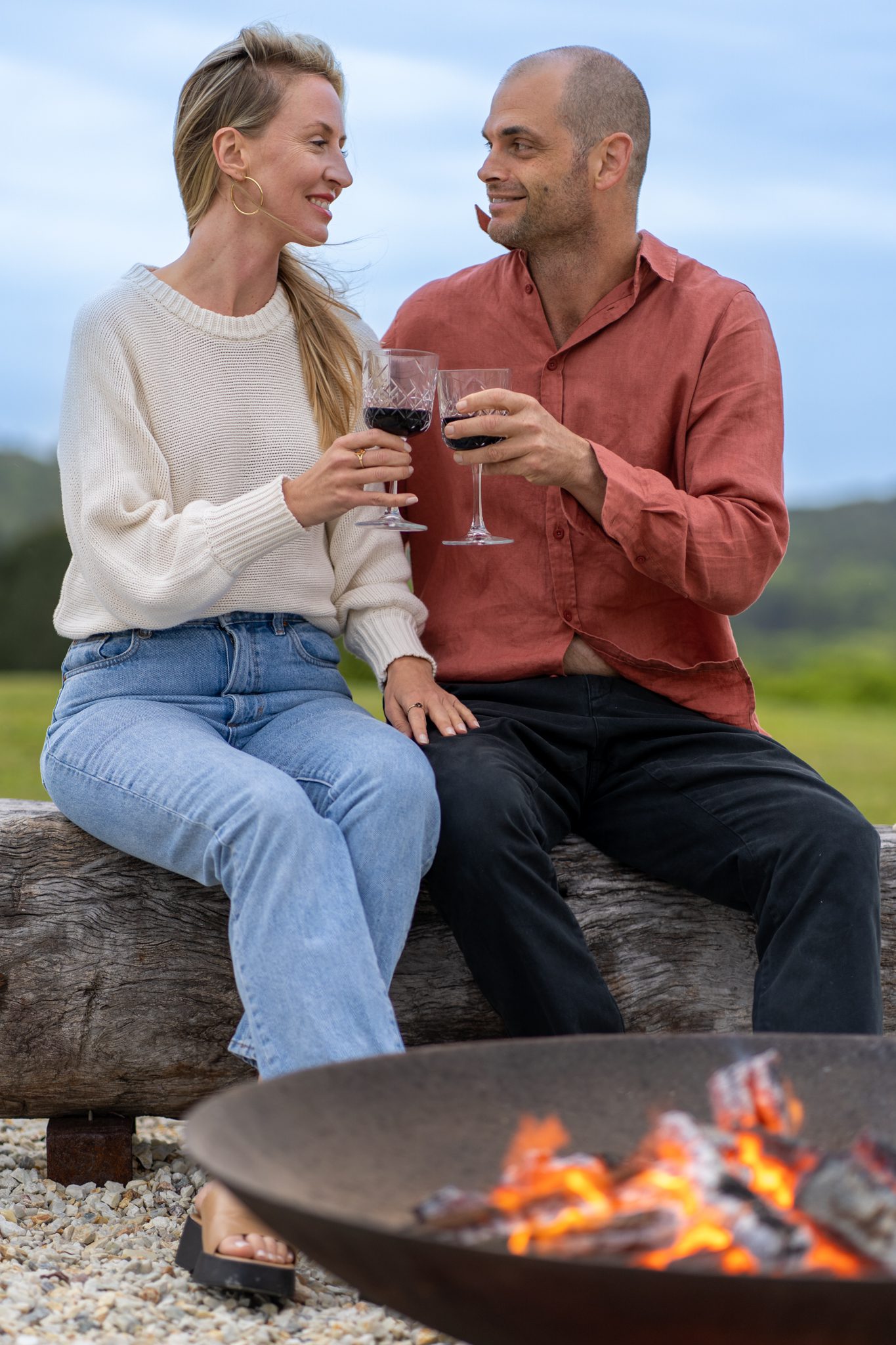 bald man and blond woman toasting red wine in front of a fire pit at heartwood farm cottage