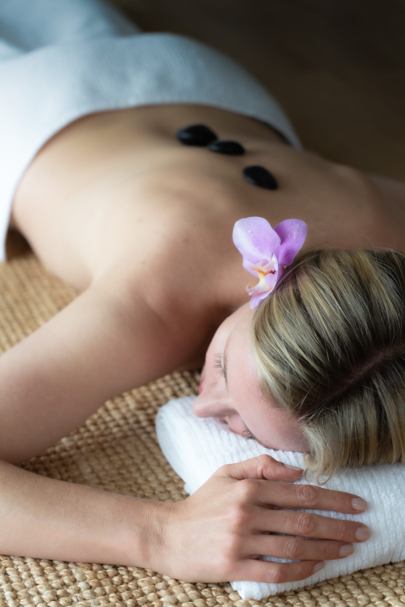 blonde woman lying facedown on a mat with a pink flower in her hair receiving a massage treatment of hot stones on her bare back at heartwood farm cottage