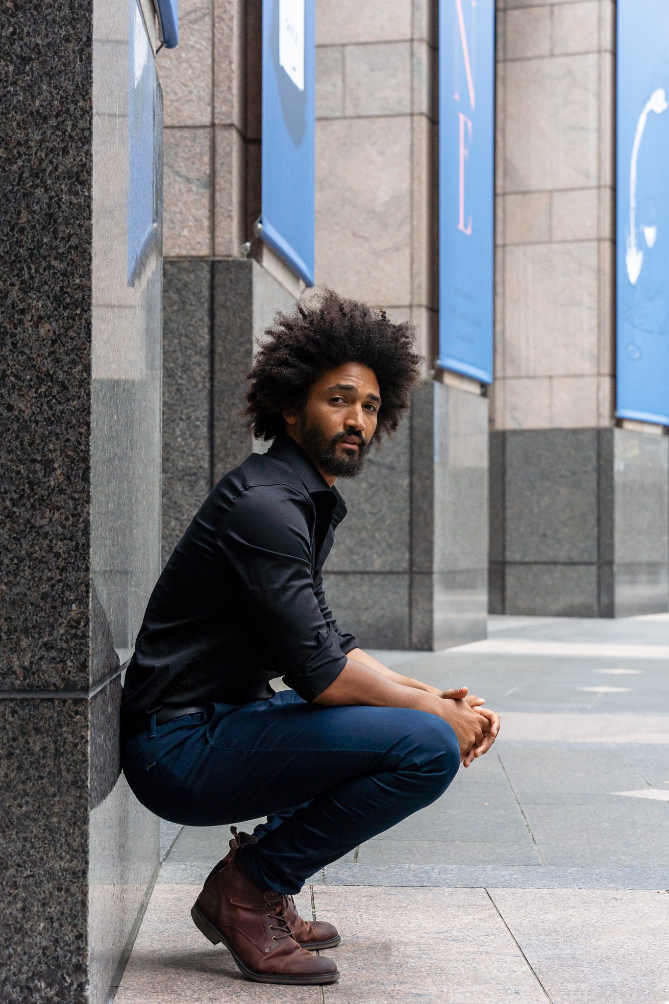 black male model crouching down next to a building pillar looking straight at the camera he is wearing black while he is photographed for the model portfolios page