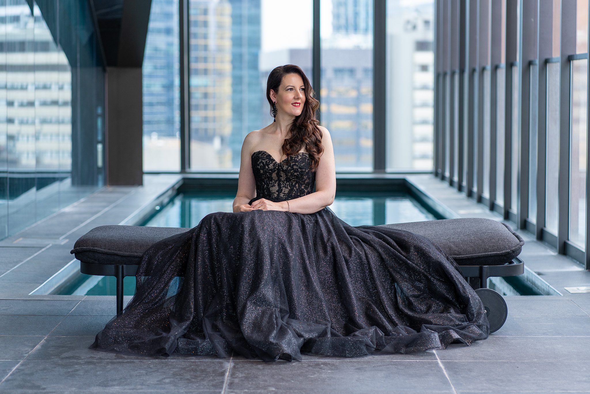 long dark-haired woman wearing a long black formal gown and sitting on a bench in a hotel pool area while she is photographed for the portraits page