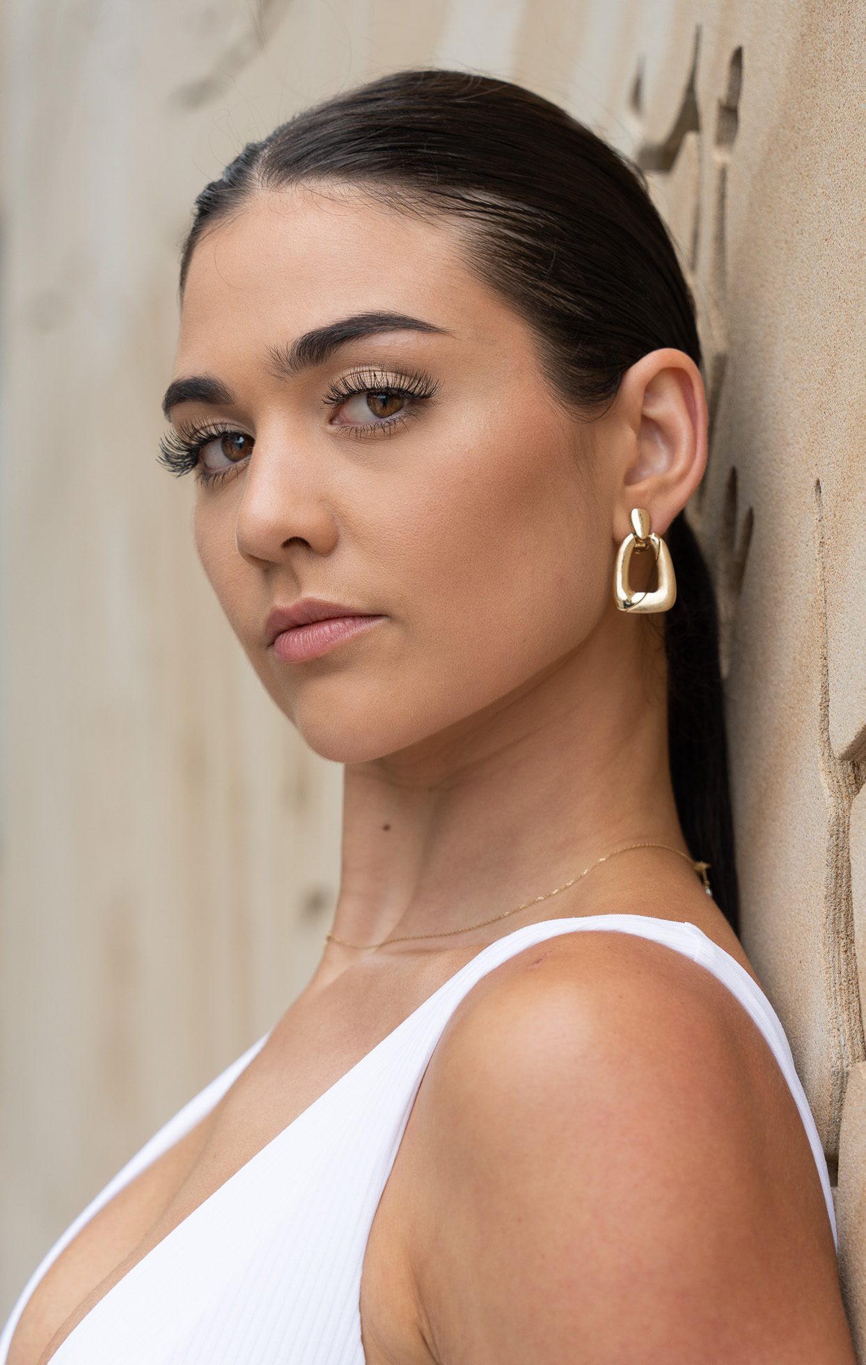 dark-haired model looking directly at the camera, wearing a white top and gold earrings standing in front of a sandstone wall and being photographed by Georgie Greene Photography for the portraits webpage