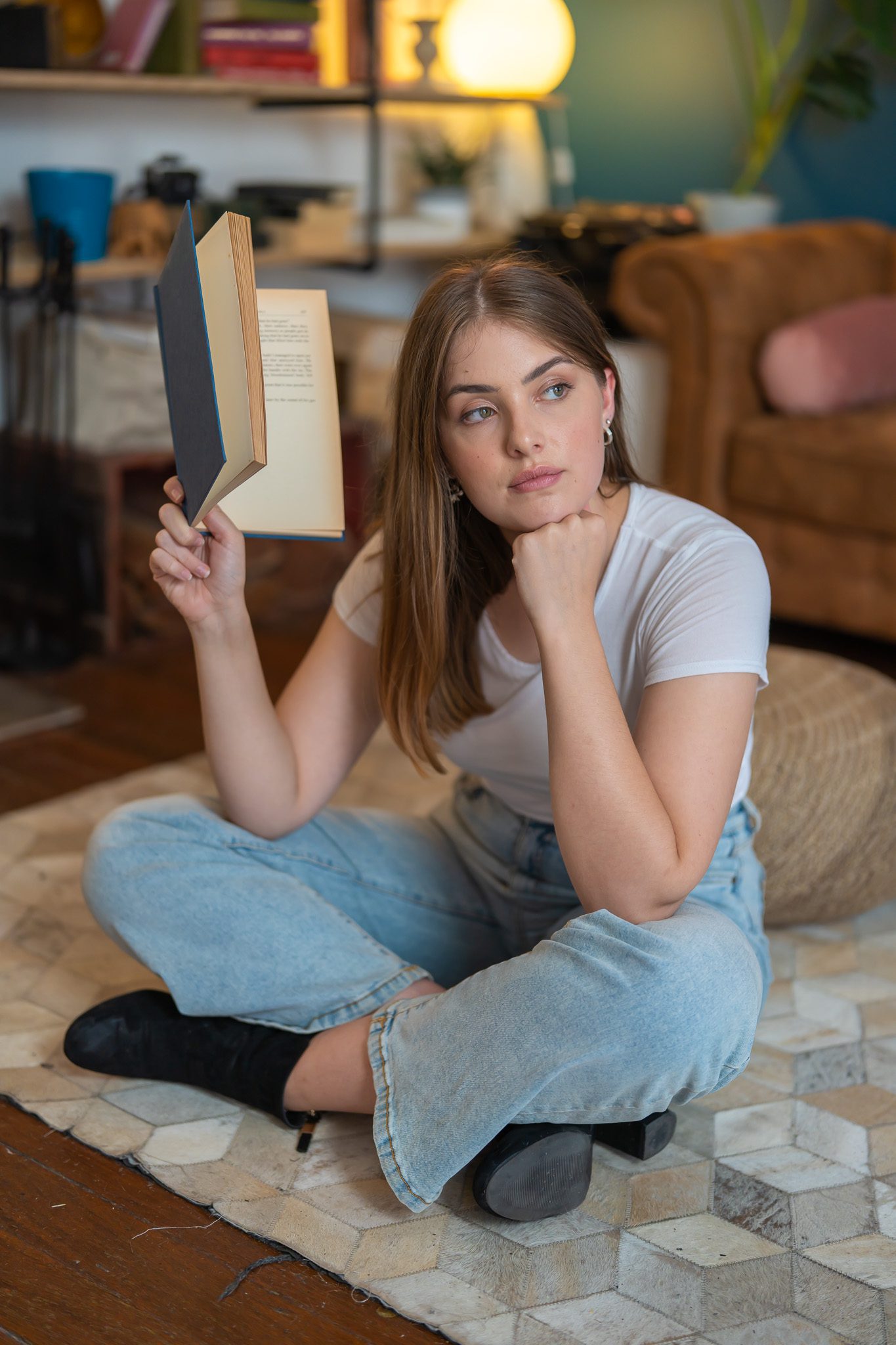 Young model sitting crossed-legged on the floor holding a book in a sitting room setting wearing a white t-shirt and jeans as she is photographed by Georgie Greene Photography for the portraits webpage