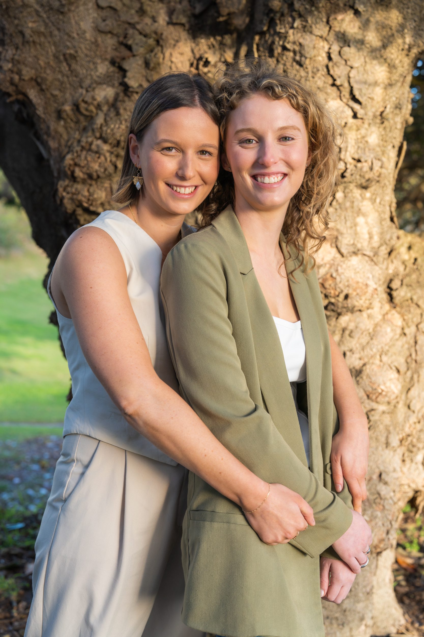 Twin sisters holding each other under a paper bark tree and smiling at the camera for the portraits webpage