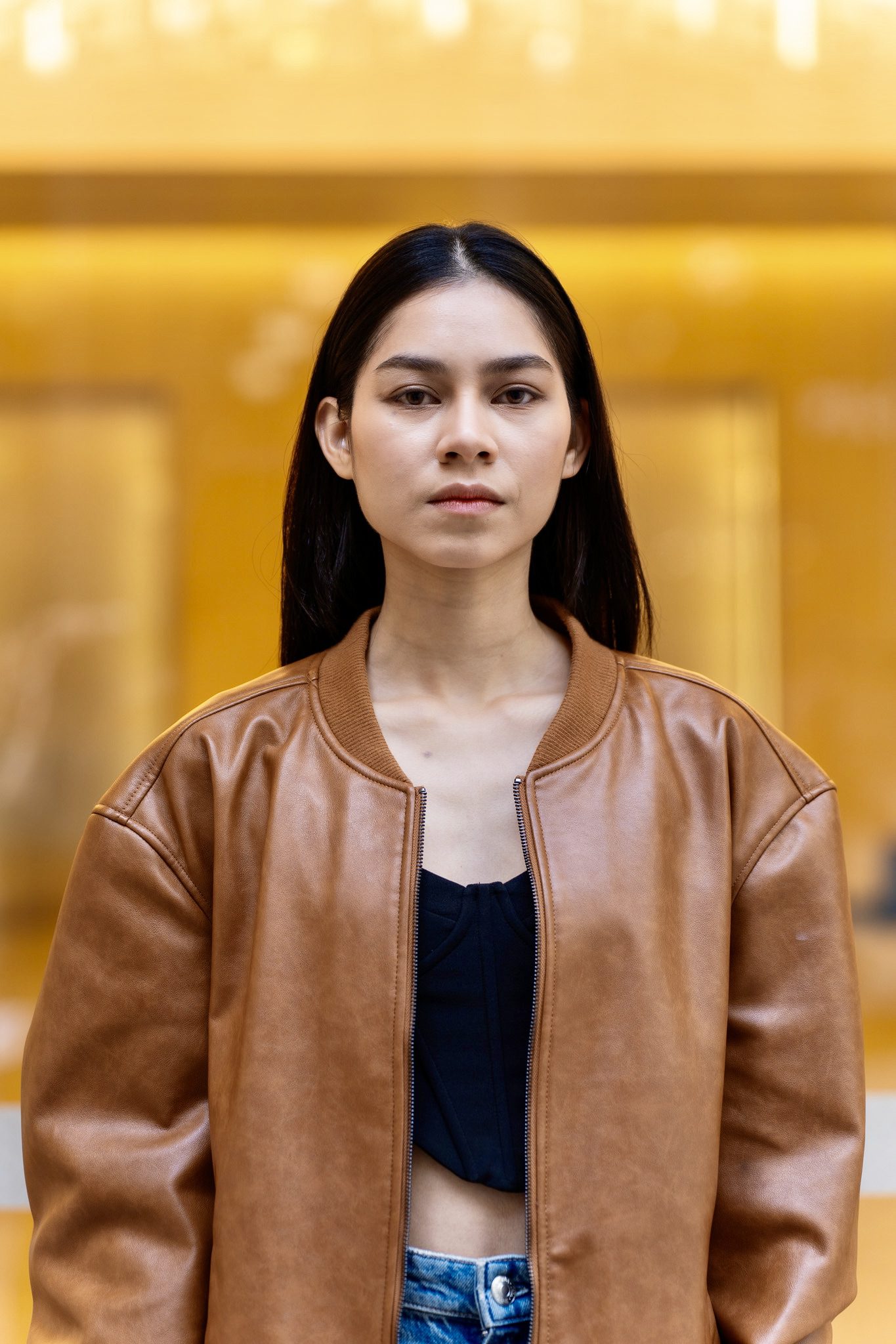 Young Asian model wearing a leather jacket and standing in front of a orange lit shopfront while here portraits are being taken
