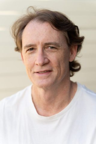 a white middle-aged man with long hair wearing a white t-shirt and looking into the camera as his corporate headshots are taken