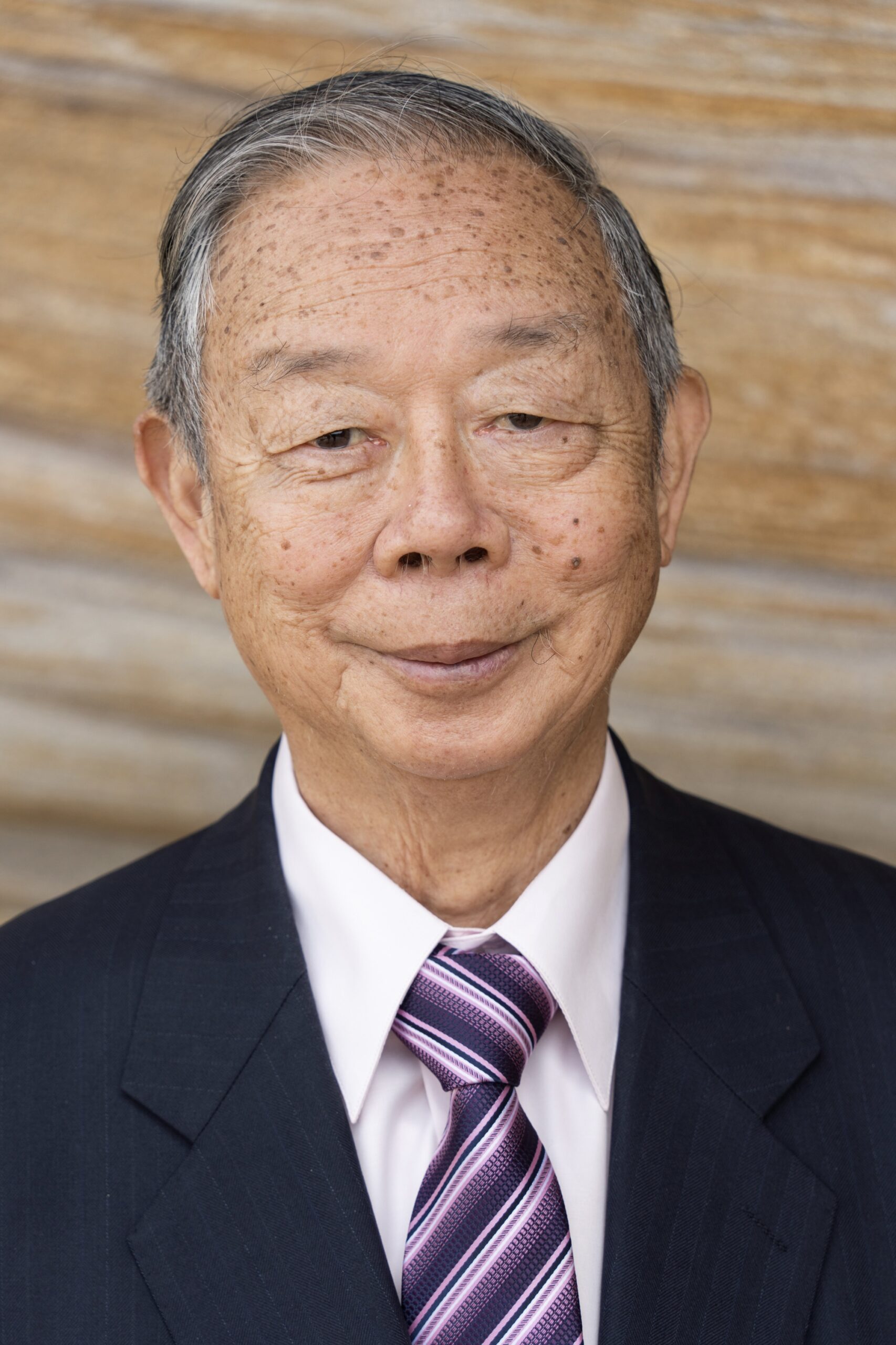 An old Asian man with short hair wearing a suit and tie and looking into the camera as his corporate headshots are taken
