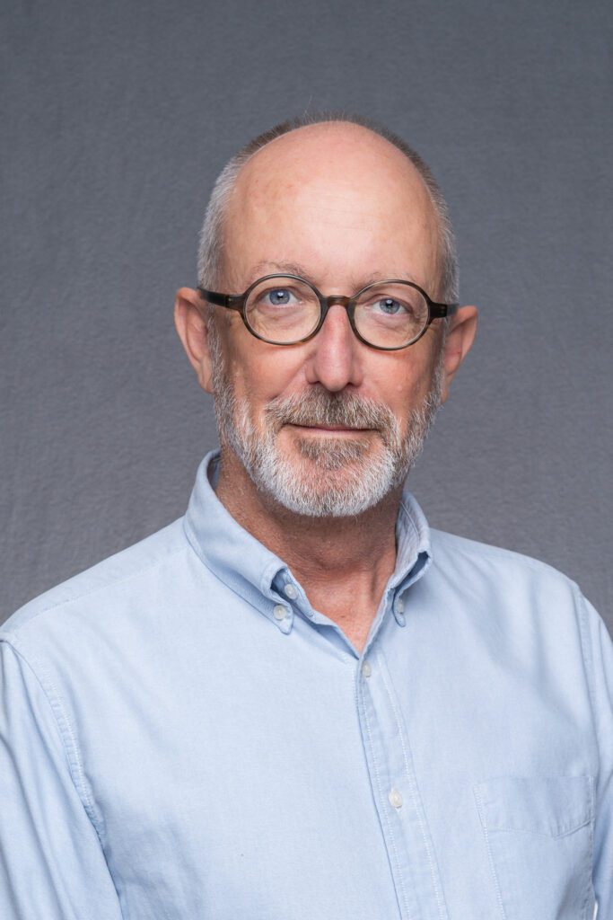 white-haired bearded man wearing glasses and a blue shirt as his blue eyes look directly into the camera as he has his professional headshot taken