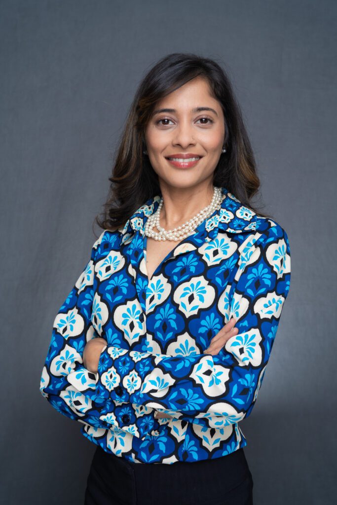 a professional Indian woman with long dark hair and brown eyes wearing a blue and white top and having her corporate headshot taken