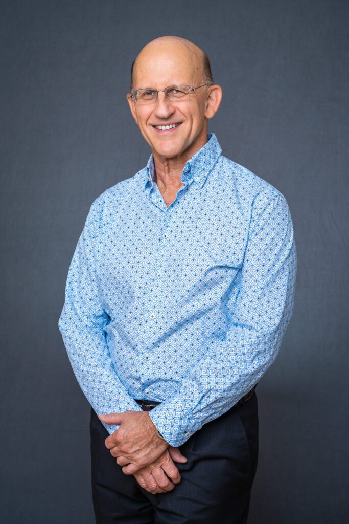 A bald older man with glasses wearing a blue shirt and having a headshot taken by Georgie Greene Photography