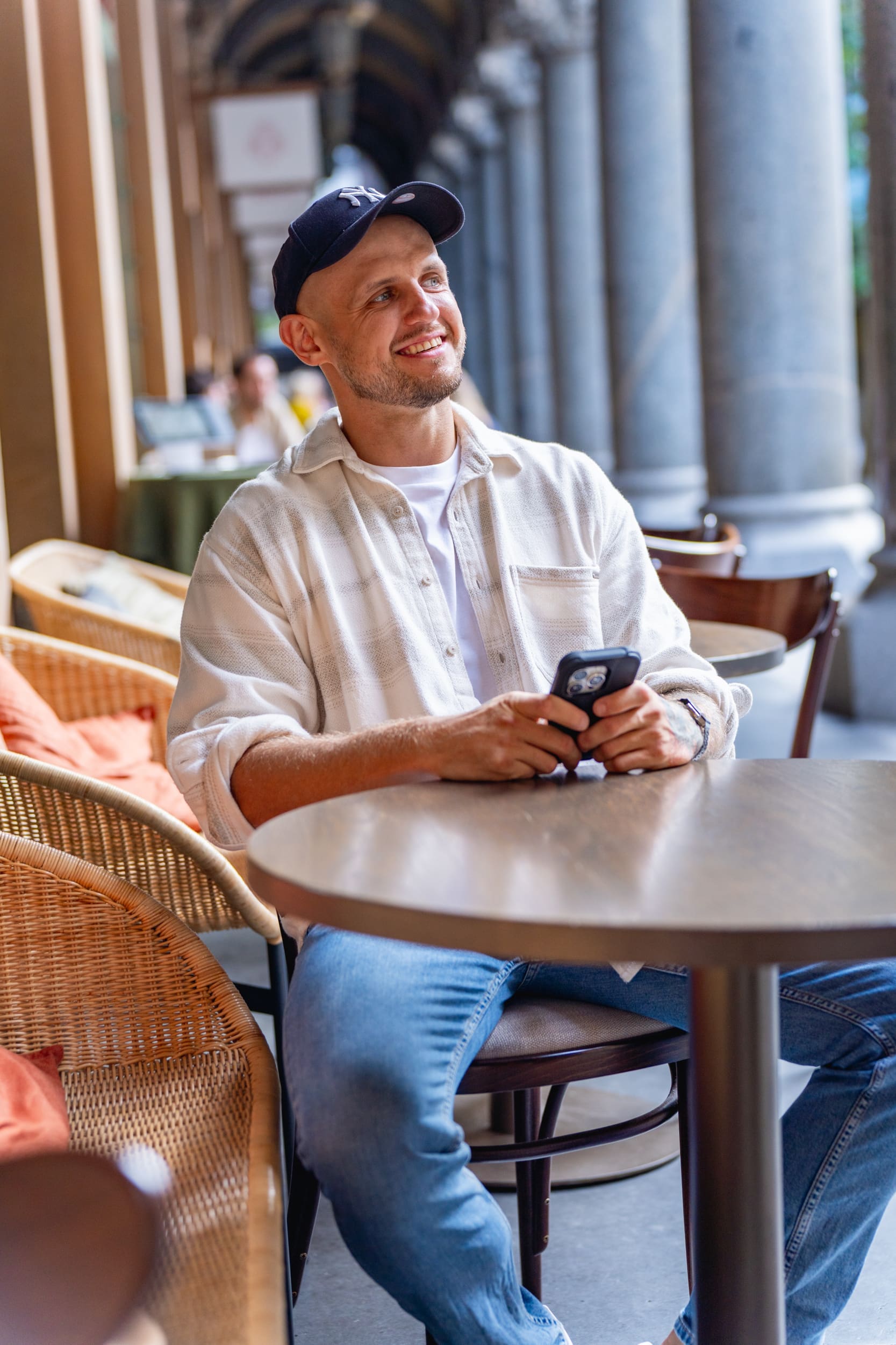 Ext: Young bald man in a white shirt sitting at a cafe table having his dating photo taken by dating photographer Georgie Green in Sydney NSW