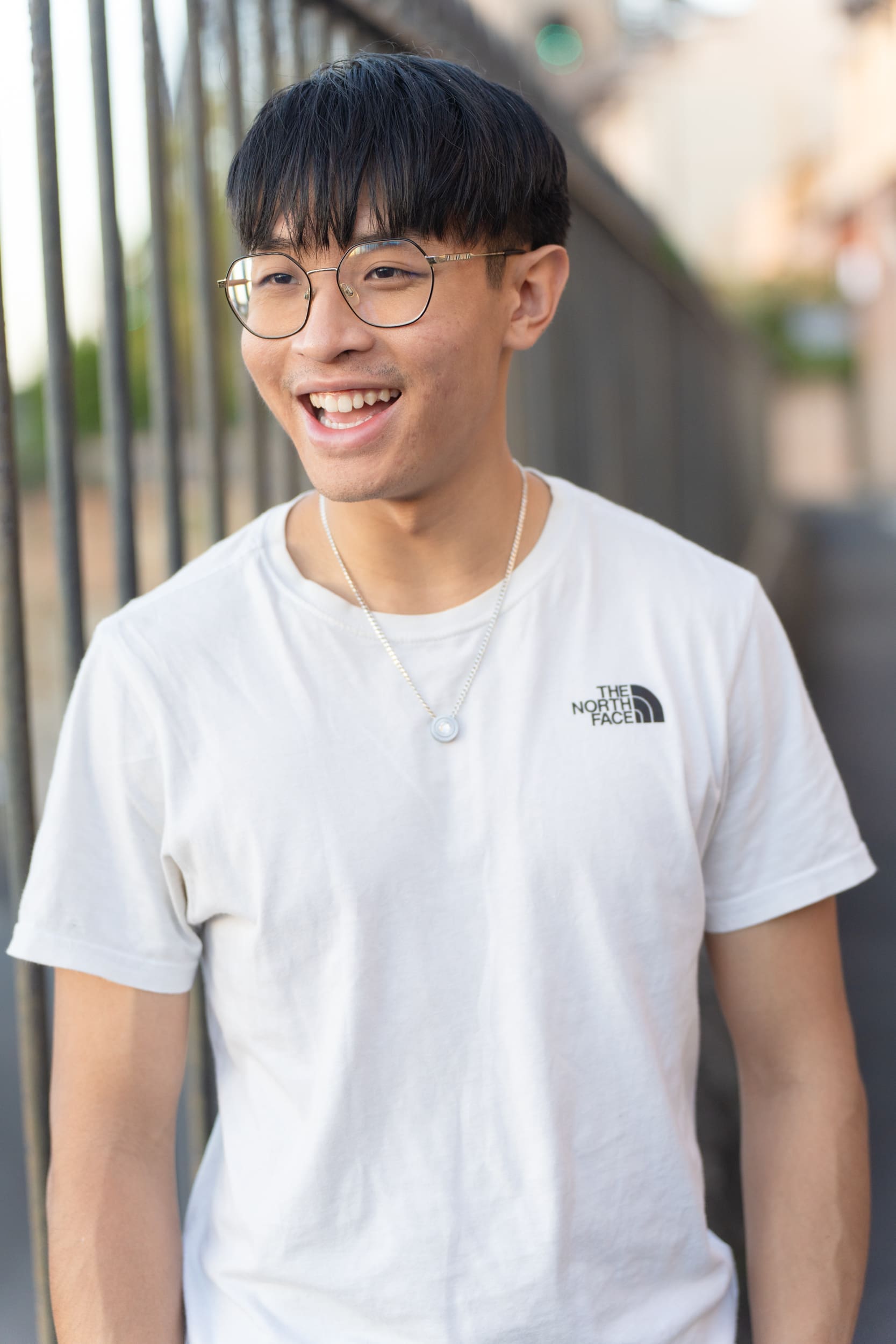 Young asian dark haired man wearing a white t-shirt and standing by a metal fence having his dating photo taken by Sydney dating photographer Georgie Greene