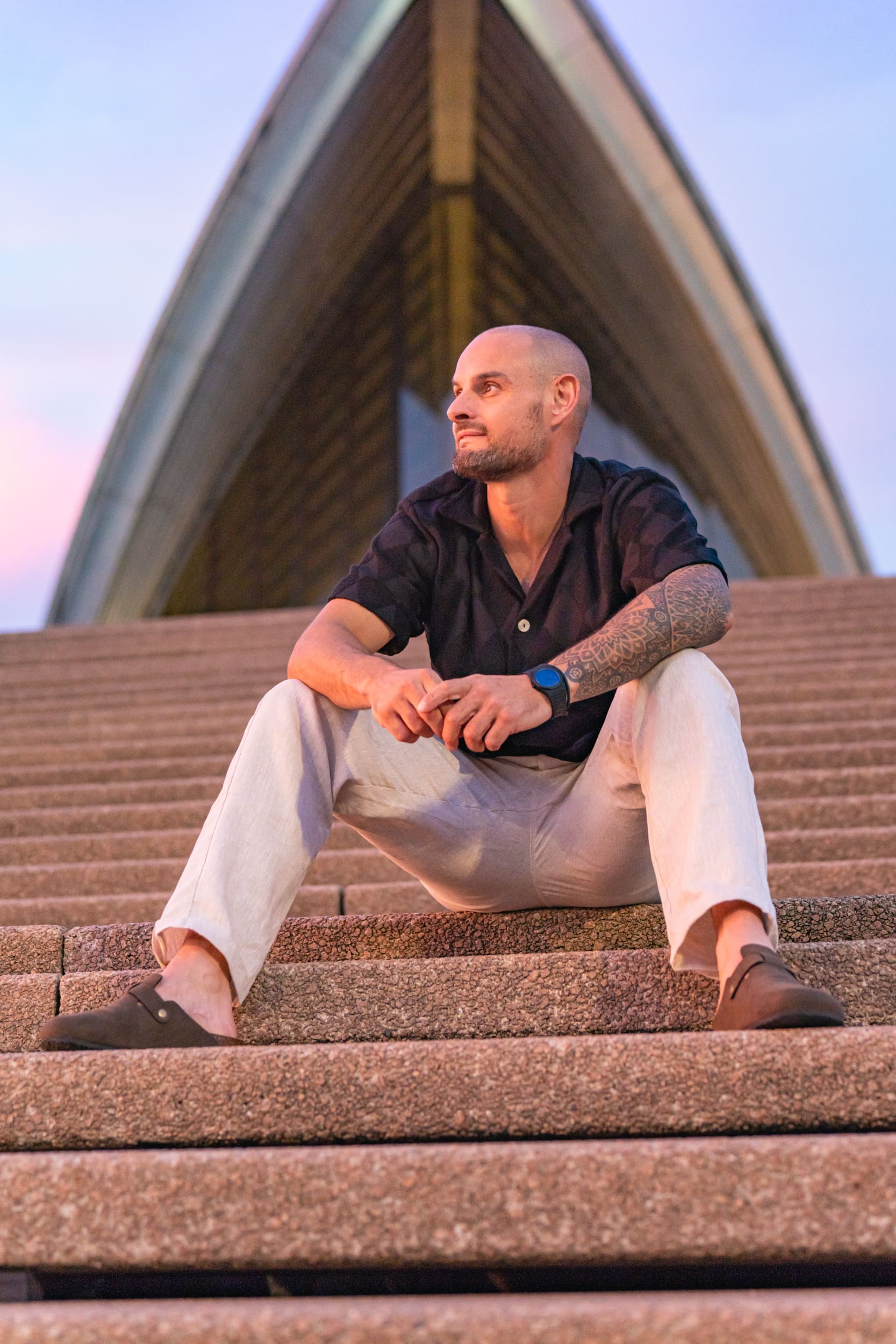 Young bald man wearing a black shirt and sitting on the steps of the Sydney Opera House having his dating photo taken by Sydney dating photographer Georgie Greene